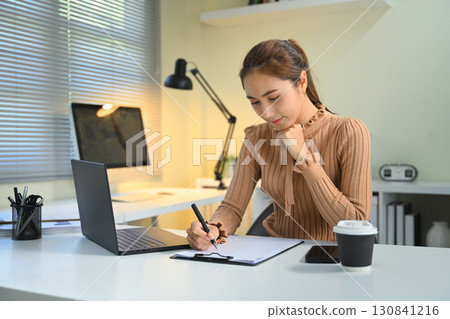 Focused young woman writing notes on clipboard while working at desk with laptop Focused young woman writing notes on clipboard while working at desk with laptop 130841216