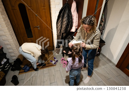Joyful moments in a cozy entryway filled with family warmth as a mother prepares her daughter while another child explores their footwear collection during a chilly evening Joyful moments in a cozy entryway filled with family warmth as a mother prepares her daughter while another child explores their footwear collection during a chilly evening 130841238