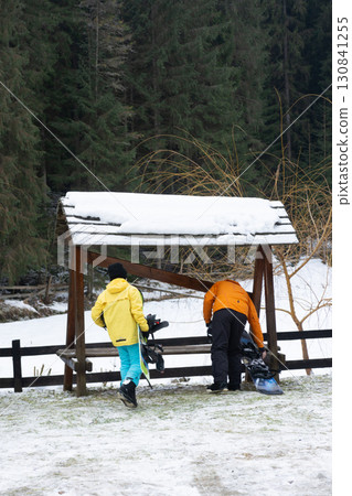 Winter adventure begins as two friends prepare to enjoy a snowy landscape surrounded by towering trees near a rustic shelter Winter adventure begins as two friends prepare to enjoy a snowy landscape surrounded by towering trees near a rustic shelter 130841255