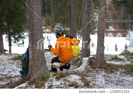 Skiers enjoying the winter chill while resting on a swing beneath tall fir trees in a snowy landscape during a vibrant day at the ski resort Skiers enjoying the winter chill while resting on a swing beneath tall fir trees in a snowy landscape during a vibrant day at the ski resort 130841261