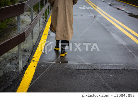 A person in a tan coat walks along a wet, yellow-lined road on a rainy day seen from behind. A woman wearing black boots with yellow soles on a street 130845892
