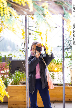 A woman capturing a moment with a camera in front of a mirror, surrounded by yellow mimosa flowers. A young girl takes a picture of herself reflection 130845895