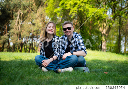 Happy Father And Daughter Sit On The Grass In The Park, Looking Into The Camera And Enjoying The Outdoors 130845954