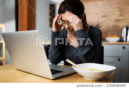 Woman Thinking About A Problem While Working On A Laptop With A Noodle Plate On The Kitchen Table 130846004