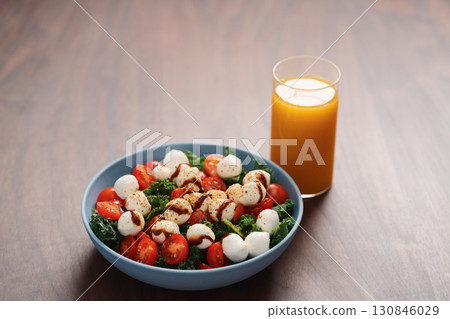 Blue bowl of salad with mozzarella, cherry tomatoes and kale, seasoned with balsamic sauce on walnut wood table with glass of orange juice 130846029
