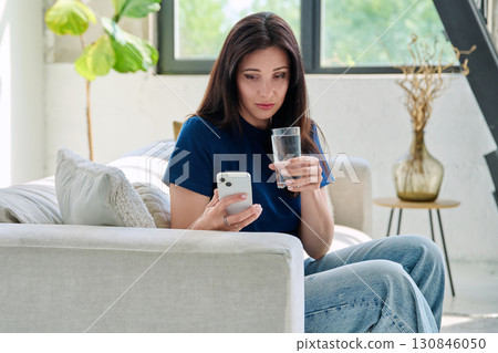 Serious sad young woman sitting at home on the couch using smartphone Serious sad young woman sitting at home on the couch using smartphone 130846050