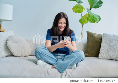 Young happy woman sitting at home on the couch using a smartphone Young happy woman sitting at home on the couch using a smartphone 130846057