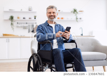Mature Caucasian man seated in wheelchair holding smartphone in contemporary home interior showcasing daily lifestyle moments 130847601