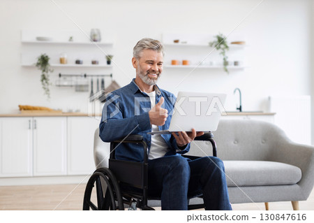 Mature Caucasian man sitting in wheelchair in home office using laptop for video call. Bright well-furnished interior creating professional yet approachable workspace environment Mature Caucasian man sitting in wheelchair in home office using laptop for video call. Bright well-furnished interior creating professional yet approachable workspace environment 130847616