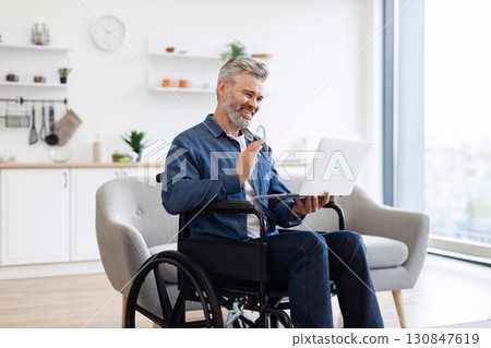 Mature Caucasian man using laptop in wheelchair having video conversation in bright home. Laptop in hands, engaged in communication. Wheelchair in modern interior showcasing accessibility 130847619