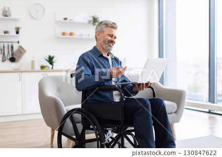 Mature Caucasian man using laptop in wheelchair having video conversation in bright home. Laptop in hands, engaged in communication. Wheelchair in modern interior showcasing accessibility 130847622