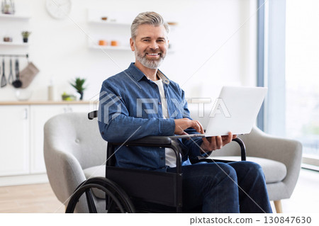 Mature Caucasian man sitting in wheelchair using laptop in bright spacious interior. Scene shows productivity, determination, and inclusivity in modern environment. 130847630