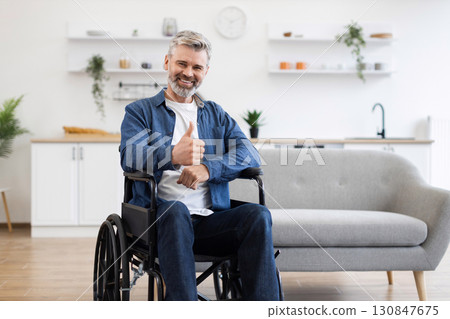 Senior Caucasian man seated in wheelchair showing thumbs-up gesture expressing positivity indoors. Image showcases accessibility, mobility aid, and cheerful demeanor in modern home environment. 130847675