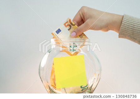 Female hand putting 50 euro banknote into glass jar, empty sticky note for inscription on it. Concept of savings Female hand putting 50 euro banknote into glass jar, empty sticky note for inscription on it. Concept of savings 130848736
