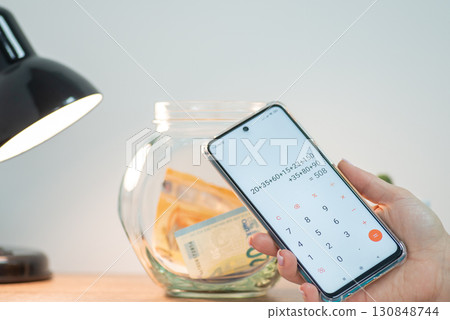 Close up of phone in female hand counting savings. Jar with euro money on the background Close up of phone in female hand counting savings. Jar with euro money on the background 130848744