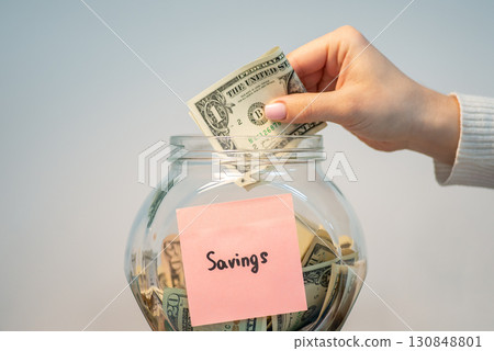 Putting one dollar banknote to glass jar with inscription savings. Concept of saving money Putting one dollar banknote to glass jar with inscription savings. Concept of saving money 130848801