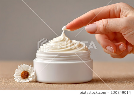 A finger gently touches rich, smooth white cream inside a jar, promoting skin beauty and natural care with a fresh daisy. 130849014