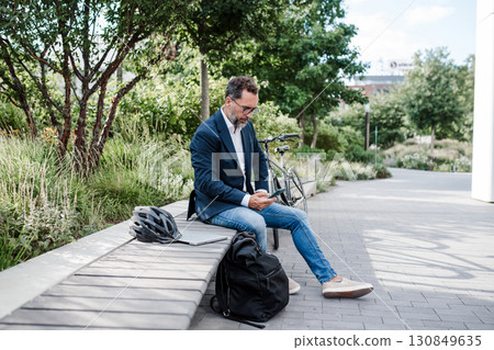 Business commuter taking a break with smartphone and bicycle in urban park. 130849635