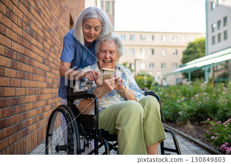 Senior patient in wheelchair showing picture in smartphone to caregiver. Senior patient in wheelchair showing picture in smartphone to caregiver. 130849803