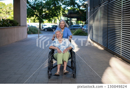 Older nurse pushing senior woman in wheelchair and talking with her. Older nurse pushing senior woman in wheelchair and talking with her. 130849824