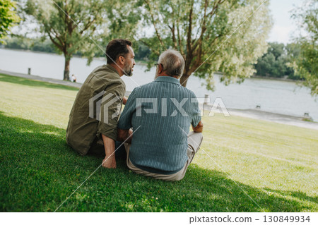 Father and adult son sitting on grass in park, sharing peaceful moment. 130849934