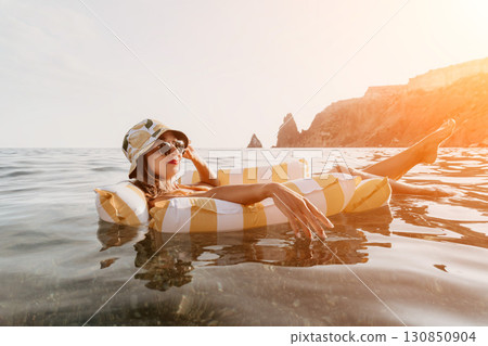 Woman Beach Float Relaxation - A woman wearing a hat and sunglasses relaxes on a striped inflatable float in the ocean near a rocky coastline. Woman Beach Float Relaxation - A woman wearing a hat and sunglasses relaxes on a striped inflatable float in the ocean near a rocky coastline. 130850904