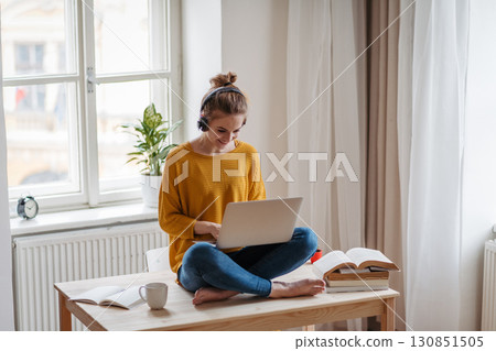 Young female student sitting at the table, using headphones while studying. 130851505