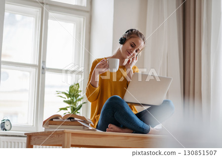 Young female student sitting at the table, using headphones while studying. 130851507