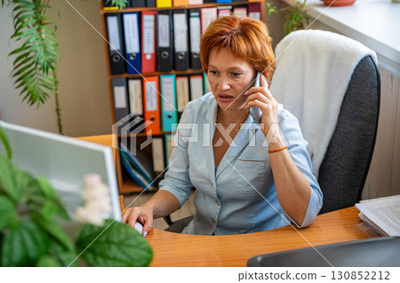Woman sitting at her desk in the office, talking on the phone while working on the computer. Business communication, multitasking, and everyday coordination at the workplace. Woman sitting at her desk in the office, talking on the phone while working on the computer. Business communication, multitasking, and everyday coordination at the workplace. 130852212