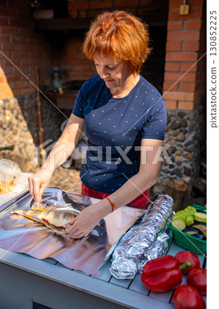 Woman seasoning fish and wrapping it in foil for outdoor grilling in her backyard. Homemade cooking, natural ingredients, and traditional barbecue preparation. 130852225