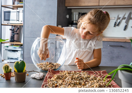 Little girl pouring stones into glass container to create drainage for future florarium or closed ecosystem. Hands-on learning, eco-friendly creativity and early involvement in nature-based projects. Little girl pouring stones into glass container to create drainage for future florarium or closed ecosystem. Hands-on learning, eco-friendly creativity and early involvement in nature-based projects. 130852245