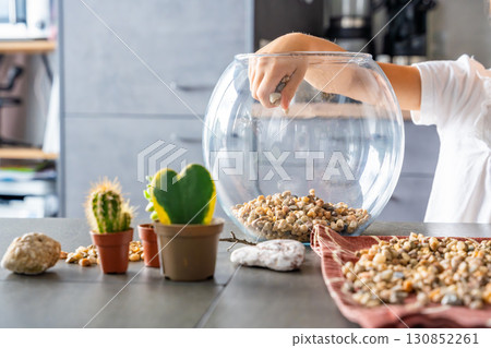 Close-up of child hands pouring stones into a glass container while preparing drainage for a florarium or closed ecosystem. Tactile learning, mindful creativity and eco-friendly crafting. Close-up of child hands pouring stones into a glass container while preparing drainage for a florarium or closed ecosystem. Tactile learning, mindful creativity and eco-friendly crafting. 130852261