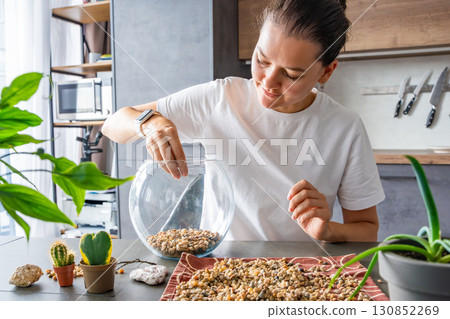 Young woman pouring stones into a glass container while preparing a terrarium. Concept of mindful crafting, eco-friendly lifestyle and home plant design. 130852269
