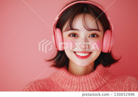 A smiling Asian girl with headphones on her head, set against a soft pink background, embodying the pleasure of listening to music or studying in a relaxed and joyful moment 130852996