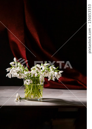 Bouquet of white snowdrops Galanthus nivalis in glass jar on dark tones with red fabric on the background Bouquet of white snowdrops Galanthus nivalis in glass jar on dark tones with red fabric on the background 130853553