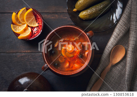 Homemade Traditional Russian soup Solyanka with meat, sausages, vegetables, pickles and olives with lemon in ceramic pot with wooden spoon on dark wooden background, 130853618