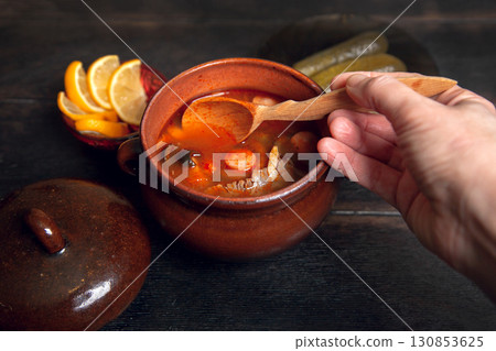 Woman scoops Homemade Traditional Russian soup Solyanka with meat, sausages, vegetables, pickles and olives with lemon in ceramic pot with wooden spoon on dark wooden background Woman scoops Homemade Traditional Russian soup Solyanka with meat, sausages, vegetables, pickles and olives with lemon in ceramic pot with wooden spoon on dark wooden background 130853625