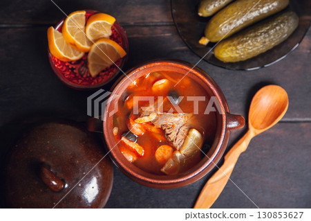 Homemade Traditional Russian soup Solyanka with meat, sausages, vegetables, pickles and olives with lemon in ceramic pot with wooden spoon on dark wooden background 130853627