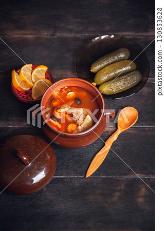 Homemade Traditional Russian soup Solyanka with meat, sausages, vegetables, pickles and olives with lemon in ceramic pot with wooden spoon on dark wooden background 130853628