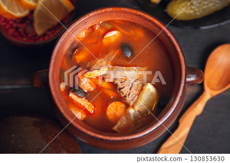 Homemade Traditional Russian soup Solyanka with meat, sausages, vegetables, pickles and olives with lemon in ceramic pot with wooden spoon on dark wooden background Homemade Traditional Russian soup Solyanka with meat, sausages, vegetables, pickles and olives with lemon in ceramic pot with wooden spoon on dark wooden background 130853630
