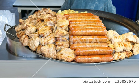 Street food scene, with a large skillet full of succulent grilled chicken pieces and plump, sizzling sausages next to golden-fried potatoes, embodying the spirit of quick, delicious outdoor dining 130853679