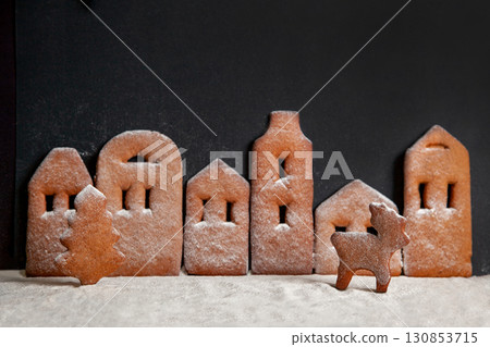 Homemade gingerbread town standing in row with Christmas tree and deer sprinkled with powdered sugar on black background Homemade gingerbread town standing in row with Christmas tree and deer sprinkled with powdered sugar on black background 130853715