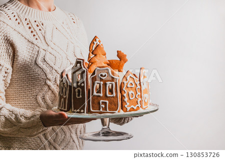 Woman holds homemade cake with gingerbread city house, Christmas tree and reindeer on glass stand Woman holds homemade cake with gingerbread city house, Christmas tree and reindeer on glass stand 130853726
