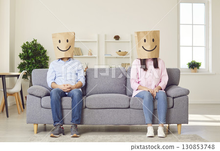 Happy anonymous couple wearing smiley face paper bags on heads sitting on sofa at home Happy anonymous couple wearing smiley face paper bags on heads sitting on sofa at home 130853748