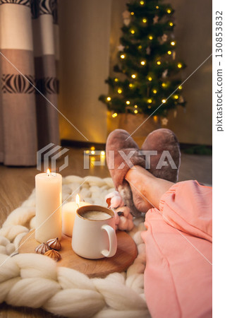 Women's feet in home fur slippers, cup of cappuccino, candles on blanket of thick yarn and Christmas tree with lights. 130853832