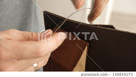 Close-up of sewing brown leather edge with thread during handmade bag crafting process Close-up of sewing brown leather edge with thread during handmade bag crafting process 130854173