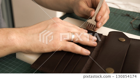 Punching stitching holes in brown leather wallet with pricking iron in artisan workshop Punching stitching holes in brown leather wallet with pricking iron in artisan workshop 130854174