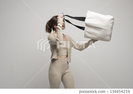 Young woman in beige clothes looking inside large white eco tote bag on grey background 130854259