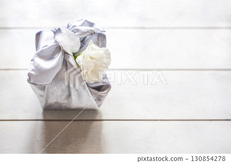 Gift box trendy wrapped in silk fabric in Furoshiki technique, white flower Eustoma or Lisianthus on light wooden background. 130854278