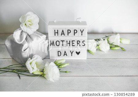 Light box with lettering Happy Mother's Day, Gift box trendy wrapped in silk fabric in Furoshiki technique, white flowers Eustoma or Lisianthus on light wooden background with copy space. 130854289